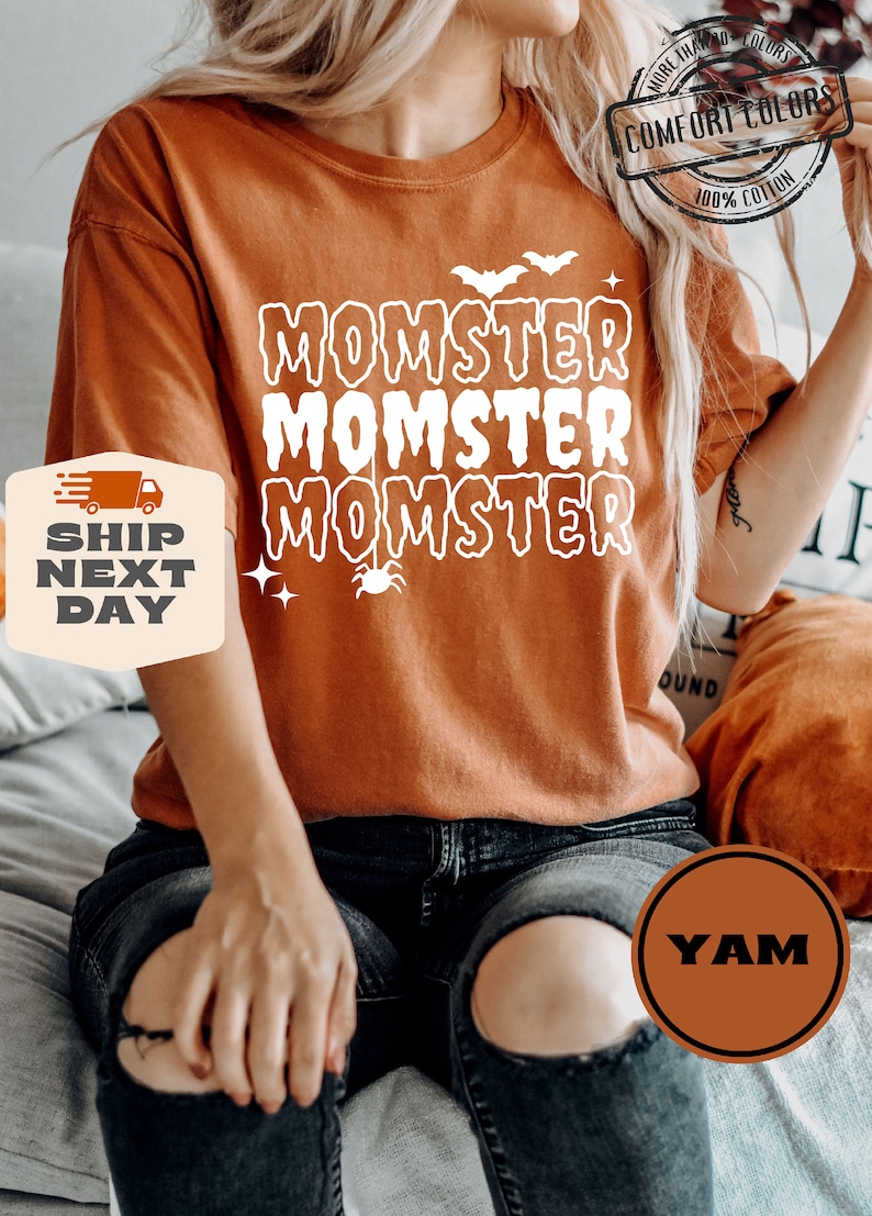 May include: A burnt orange t-shirt with the text "MOMSTER" repeated three times in white. A small spider with a web is hanging below the text. The shirt is on a person wearing black jeans.