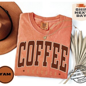 May include: A peach-colored t-shirt with the word "COFFEE" printed in brown letters. The shirt has a distressed look and is made of 100% cotton.