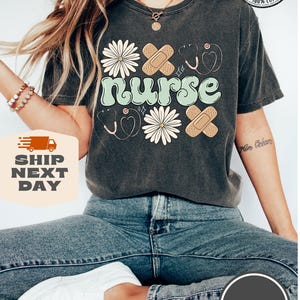 May include: A dark grey t-shirt with the word "nurse" in a retro font, surrounded by daisies, plasters, and a stethoscope. The shirt has a distressed look and is made of 100% cotton.