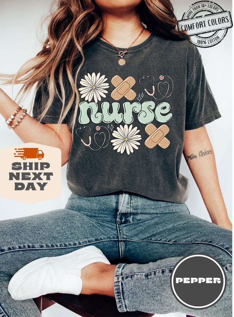 May include: A dark grey t-shirt with the word "nurse" in a retro font, surrounded by daisies, plasters, and a stethoscope. The shirt has a distressed look and is made of 100% cotton.
