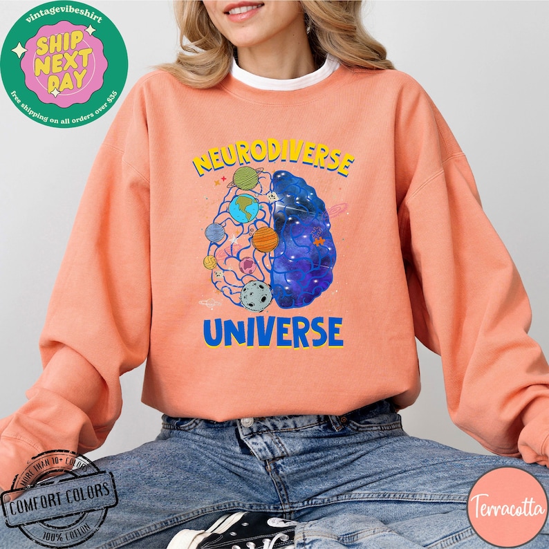 Op de afbeelding: Een terracotta oranje sweatshirt met een afbeelding van een brein met planeten en sterren erin. De tekst "Neurodiverse Universe" is op het sweatshirt gedrukt.