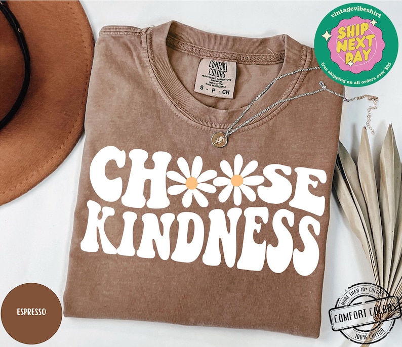 May include: A brown t-shirt with the text "Choose Kindness" in white with a daisy graphic. The shirt is made of 100% cotton.