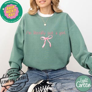 Puede incluir: Una sudadera de color verde claro con el texto "I'm literally just a girl" en letras rosas. Un lazo rosa est&aacute; centrado debajo del texto. La sudadera est&aacute; hecha de 100% algod&oacute;n.