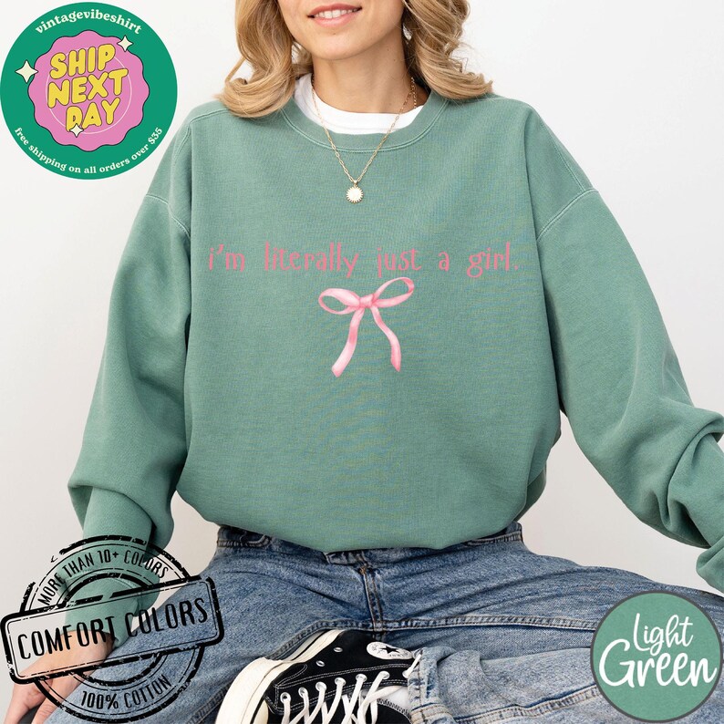 Puede incluir: Una sudadera de color verde claro con el texto "I'm literally just a girl" en letras rosas. Un lazo rosa est&aacute; centrado debajo del texto. La sudadera est&aacute; hecha de 100% algod&oacute;n.