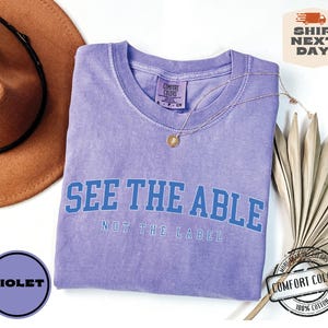 May include: Purple t-shirt with the text "See the Able, Not the Label" in blue lettering. The shirt is made of 100% cotton.