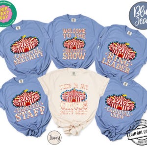 May include: A collection of blue and ivory t-shirts with circus-themed designs. The shirts feature text like "Carnival Security," "Welcome to the Greatest Show," and "This is My Circus Party Shirt." The designs include a red and white circus tent.