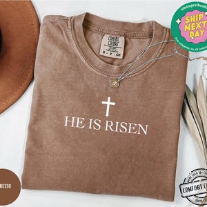 May include: A brown t-shirt with the text "HE IS RISEN" in white lettering and a white cross above the text. The shirt is made of 100% cotton.
