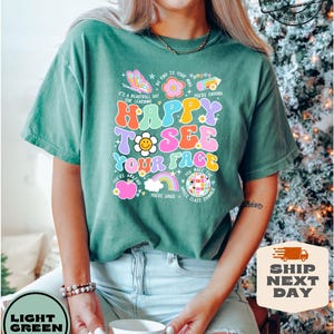 May include: A green t-shirt with a colorful graphic that says "Happy to see your face". The graphic includes a rainbow, a smiley face, an apple, and a disco ball. The text "Be kind to your mind", "It's a beautiful day for learning", "You're enough", "You're amazing", "You're loved", and "You make the whole class shimmer" is also included in the graphic.