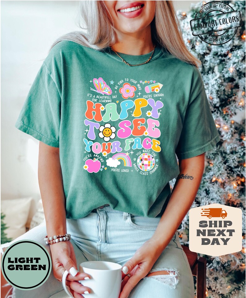 May include: A green t-shirt with a colorful graphic that says "Happy to see your face". The graphic includes a rainbow, a smiley face, an apple, and a disco ball. The text "Be kind to your mind", "It's a beautiful day for learning", "You're enough", "You're amazing", "You're loved", and "You make the whole class shimmer" is also included in the graphic.
