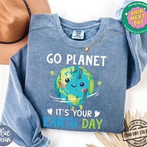 May include: A blue sweatshirt with a cartoon image of the Earth smiling and holding a sign that says "Go Planet It's Your Earth Day".