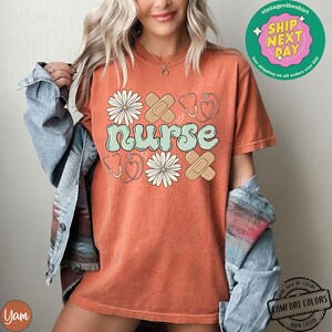 May include: A burnt orange t-shirt featuring a graphic design of the word "nurse" in a light green font. The graphic also includes a stethoscope, a heart, plasters, and daisies.