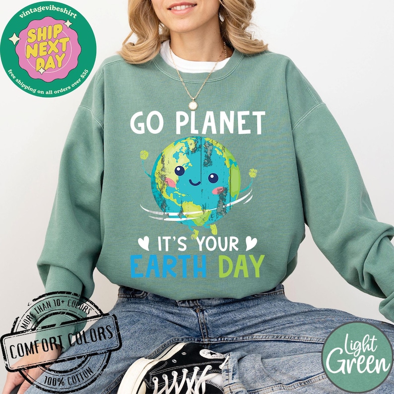 May include: A light green sweatshirt with the text "Go Planet It's Your Earth Day" and a cartoon image of a smiling Earth. The Earth is blue and green with continents and a white cloud. The text is in white, blue, and green.