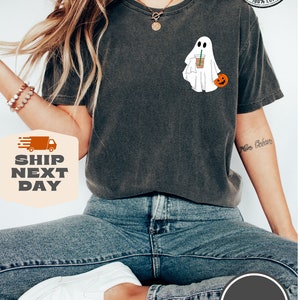 Peut inclure: Un t-shirt gris fonc&eacute; avec un fant&ocirc;me blanc tenant une tasse de caf&eacute; et une petite citrouille. Le t-shirt porte l'inscription "COMFORT COLORS" et "100% COTTON".