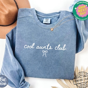 May include: A blue sweatshirt with the text "cool aunts club" in white lettering and a small white bow underneath. The sweatshirt is made of a soft, comfortable fabric.
