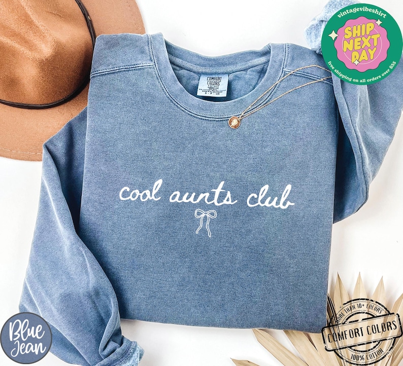 May include: A blue sweatshirt with the text "cool aunts club" in white lettering and a small white bow underneath. The sweatshirt is made of a soft, comfortable fabric.