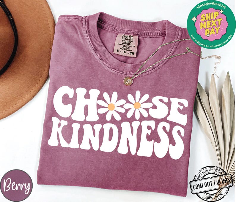 May include: A mauve colored t-shirt with the text "Choose Kindness" in white with two white daisies. The daisies have yellow centers.