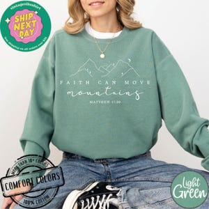 May include: A light green sweatshirt with the text "FAITH CAN MOVE MOUNTAINS" and the scripture reference "MATTHEW 17:20". The sweatshirt features a simple line drawing of mountains and stars.