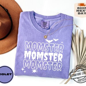 May include: A purple t-shirt with the text "Momster" repeated three times in white dripping letters. The shirt has a distressed look and is decorated with a spider and bats.