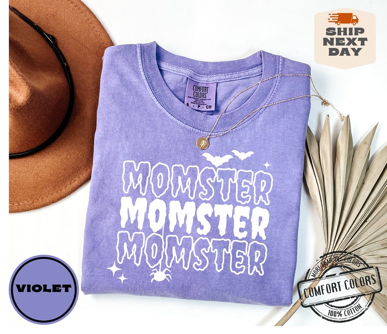 May include: A purple t-shirt with the text "Momster" repeated three times in white dripping letters. The shirt has a distressed look and is decorated with a spider and bats.