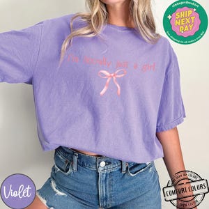 Puede incluir: Una camiseta morada con el texto "I'm literally just a girl" y un lazo rosa.