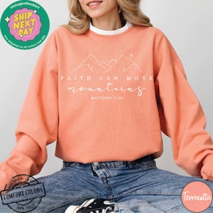 May include: A terracotta colored sweatshirt with the text "Faith can move mountains" and "Matthew 17:20" in white. The sweatshirt features a simple line drawing of mountains.