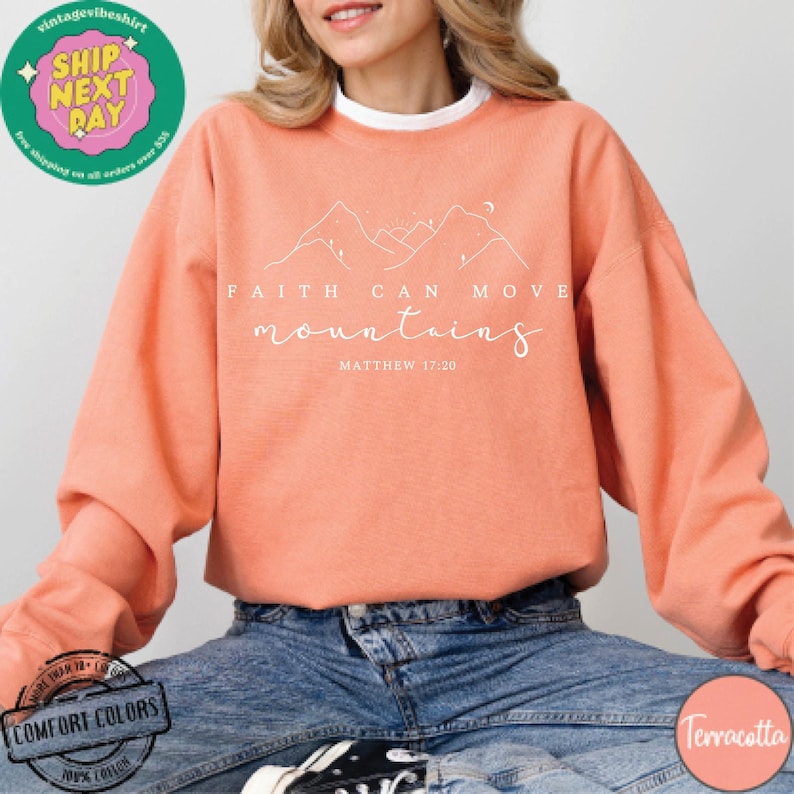 May include: A terracotta colored sweatshirt with the text "Faith can move mountains" and "Matthew 17:20" in white. The sweatshirt features a simple line drawing of mountains.