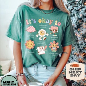 May include: A light green t-shirt with the text "It's okay to" and several cartoon illustrations. The illustrations include a brain, a flower, a heart, a flower with an X on its face, a notepad, and a flower with a sun.