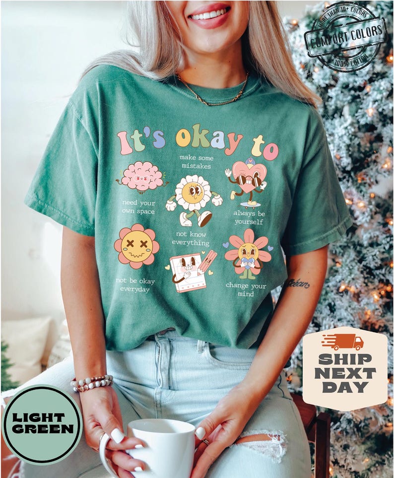 May include: A light green t-shirt with the text "It's okay to" and several cartoon illustrations. The illustrations include a brain, a flower, a heart, a flower with an X on its face, a notepad, and a flower with a sun.