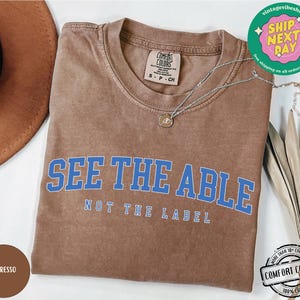May include: A brown t-shirt with the text "See the Able, Not the Label" in blue lettering. The shirt is made of 100% cotton and is available in a variety of sizes.