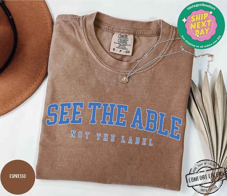 May include: A brown t-shirt with the text "See the Able, Not the Label" in blue lettering. The shirt is made of 100% cotton and is available in a variety of sizes.