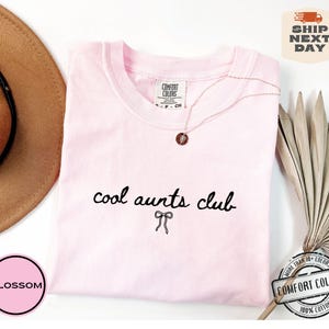 May include: Pink t-shirt with the text "cool aunts club" in black script with a small bow underneath. The shirt is on a white background with a brown hat and a dried palm leaf.