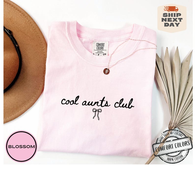 May include: Pink t-shirt with the text "cool aunts club" in black script with a small bow underneath. The shirt is on a white background with a brown hat and a dried palm leaf.