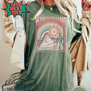 May include: Moss green Comfort Colors t-shirt with a retro graphic design. The design features a rainbow, sun, mountains, and the word "WANDERLUST". The shirt is made of 100% cotton.