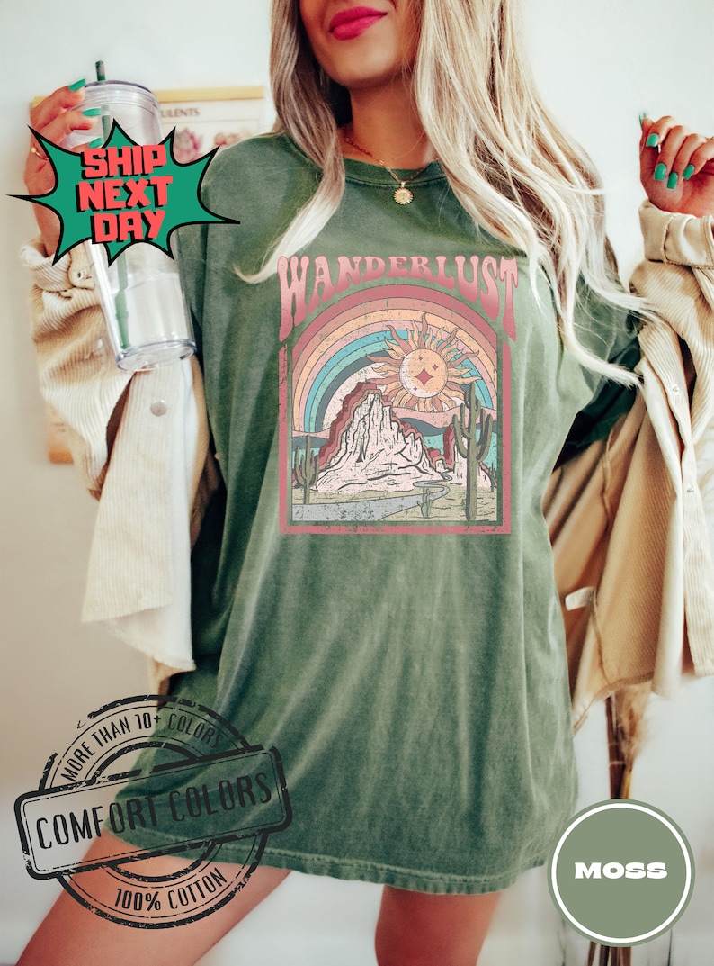 May include: Moss green Comfort Colors t-shirt with a retro graphic design. The design features a rainbow, sun, mountains, and the word "WANDERLUST". The shirt is made of 100% cotton.