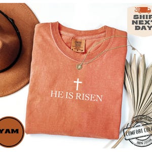 May include: A coral-colored t-shirt with a white cross and the text "HE IS RISEN". The shirt is made of 100% cotton.