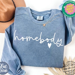 Może przedstawiać: Niebieska bluza z dżinsu z napisem "homebody" białymi literami i małym białym sercem. Bluza jest wykonana z miękkiej, wygodnej tkaniny.