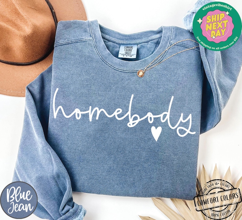 Może przedstawiać: Niebieska bluza z dżinsu z napisem "homebody" białymi literami i małym białym sercem. Bluza jest wykonana z miękkiej, wygodnej tkaniny.