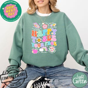 May include: Light green sweatshirt with a colorful graphic print that says "Happy to see your face". The graphic includes a rainbow, a smiley face, an apple, and the words "You're amazing", "You're loved", and "You make the whole class shimmer".