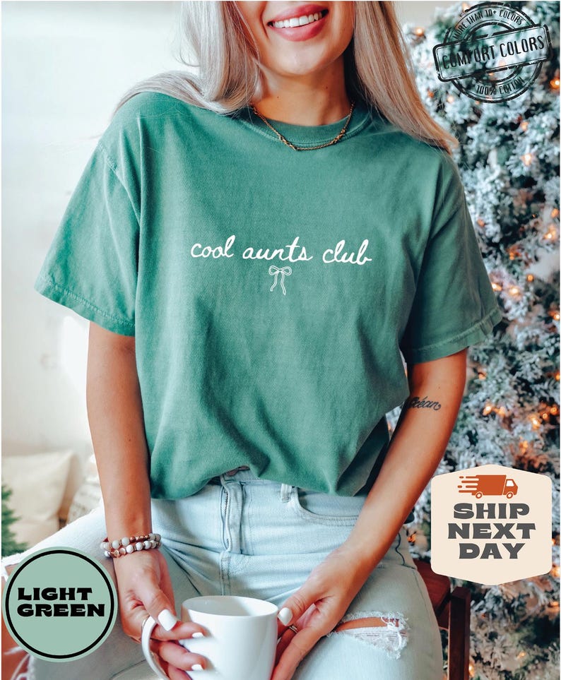 May include: A light green t-shirt with the text "cool aunts club" in white lettering. The shirt is worn by a person with a white gold necklace and a light blue denim skirt.