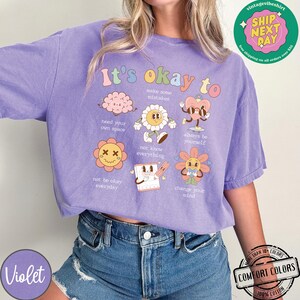 May include: Purple t-shirt with the text "It's okay to" and illustrations of flowers, a brain, a heart, and a notepad. The illustrations are in pastel colors and have a retro style. The text is in a rainbow font.