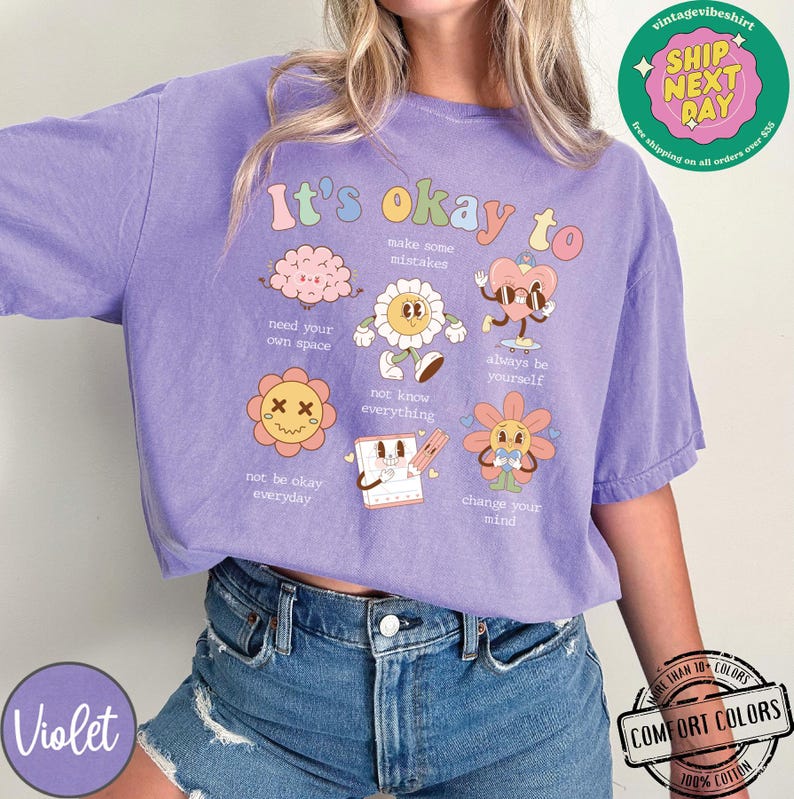 May include: Purple t-shirt with the text "It's okay to" and illustrations of flowers, a brain, a heart, and a notepad. The illustrations are in pastel colors and have a retro style. The text is in a rainbow font.