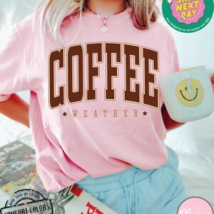 May include: A pink t-shirt with the text "COFFEE WEATHER" in brown letters with a gold outline. The shirt is being worn by a person with blonde hair and blue shorts.