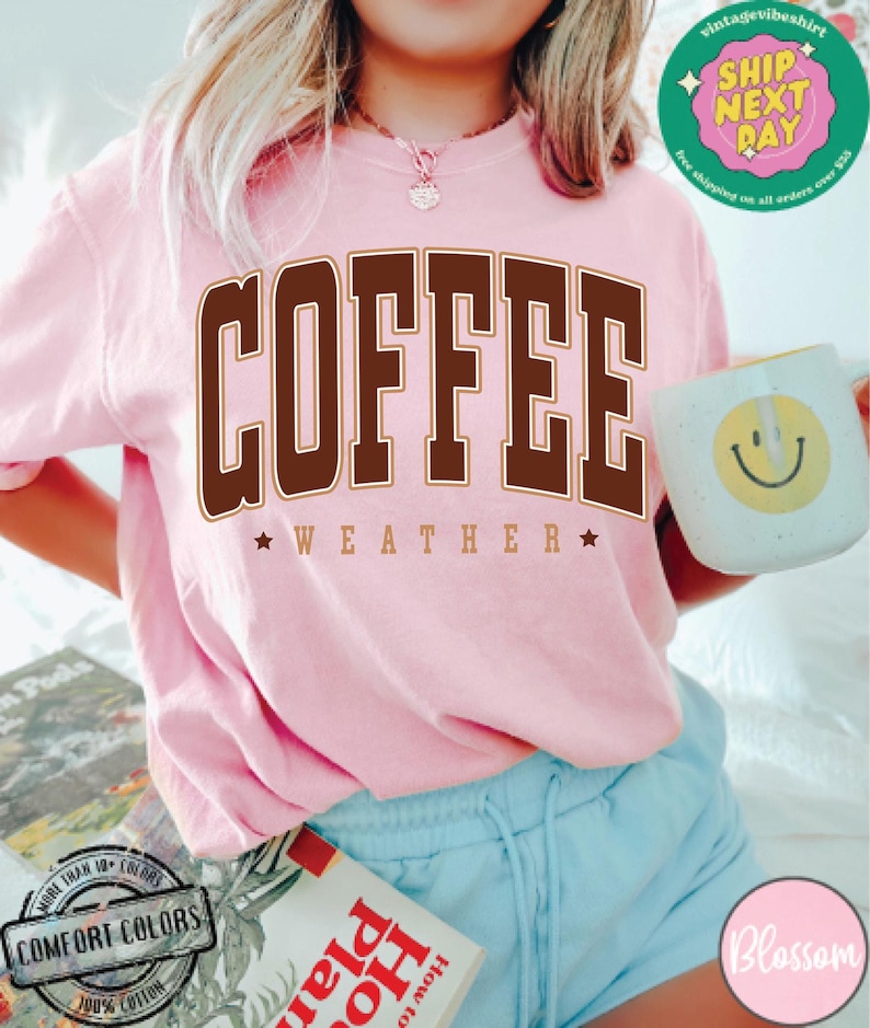 May include: A pink t-shirt with the text "COFFEE WEATHER" in brown letters with a gold outline. The shirt is being worn by a person with blonde hair and blue shorts.