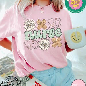 May include: A pink t-shirt with the word "nurse" in green and white lettering. The shirt is decorated with a stethoscope, a heart, bandages, and flowers.
