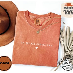May include: A peach-colored t-shirt with the text "IN MY GRANDMA ERA" and a small white heart. The shirt is made of 100% cotton.