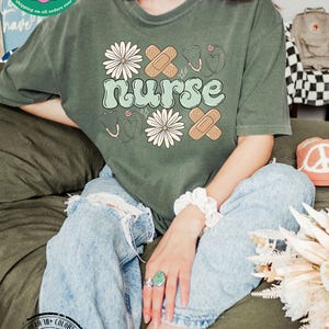 May include: A green t-shirt featuring the word "nurse" in a retro font, surrounded by white daisies, plasters, and stethoscopes. The shirt is made from 100% cotton and is available in a range of colours.
