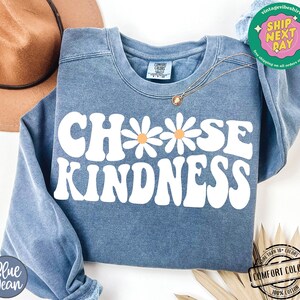 May include: A blue sweatshirt with the text "Choose Kindness" in white with a retro font. The text is surrounded by three white daisies with orange centers.