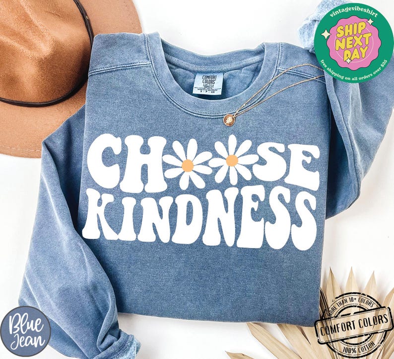 May include: A blue sweatshirt with the text "Choose Kindness" in white with a retro font. The text is surrounded by three white daisies with orange centers.