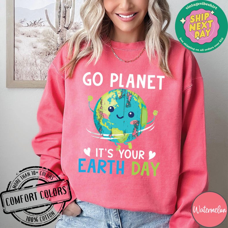 May include: Pink sweatshirt with a graphic of a smiling cartoon Earth holding a sign that says "Go Planet It's Your Earth Day".