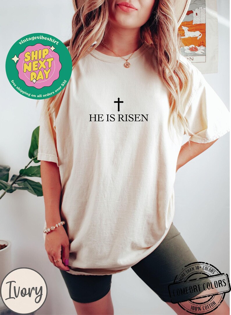 May include: A white t-shirt with the text "HE IS RISEN" and a cross above it. The shirt is being worn by a person with long blonde hair.